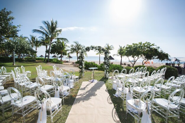 Outdoor beach wedding setup with white chairs, a floral arch, and a sandy aisle surrounded by palm trees and ocean views.