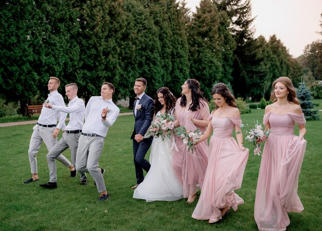 Joyful bride, groom, bridesmaids in blush pink dresses, and groomsmen in white shirts walking together on a grassy lawn.