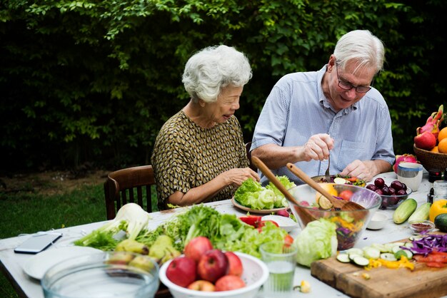 An elderly couple is preparing a fresh salad outdoors, smiling as they chop vegetables and fruit at a garden table.