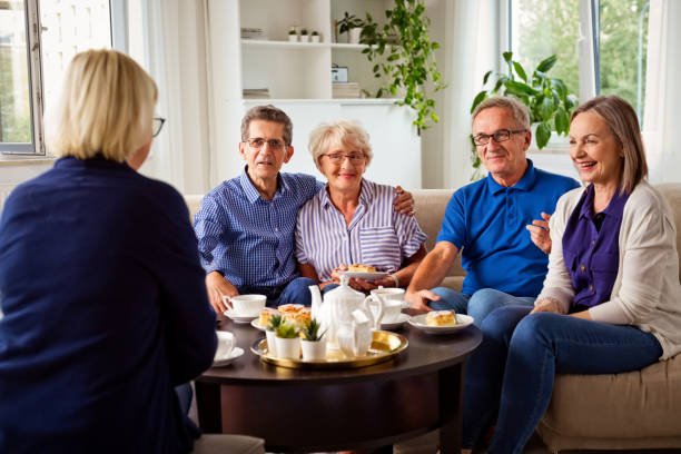 Four seniors sitting together on a sofa, smiling and chatting over coffee and dessert in a bright, cozy living room.