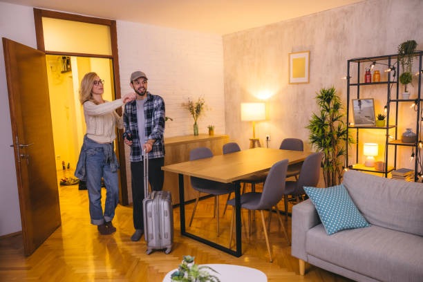 A couple entering a cozy short-term rental apartment, woman pointing around while man holds suitcase.